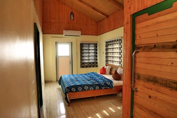 Ein Schlafzimmer mit Holzboden, einem Bett mit gemustertem Überwurf, einem Fenster mit Jalousien und einer Tür mit grünem Rahmen.