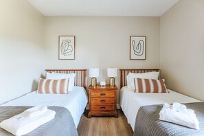 Hotellihuoneessa on kaksi sänkyä, joissa kummassakin on tyyny ja taitettu pyyhe. Sängyn vieressä on puinen yöpöytä, jossa on kaksi lamppua, ja sänkyjen välissä on pieni pöytä.