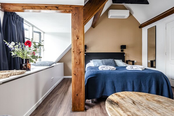 A bedroom with a wooden ceiling, a bed with a blue bedspread, a bedside table, a window with curtains, and a vase with flowers.