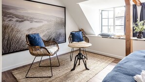 Una habitación acogedora con un cuadro de paisaje grande, dos sillones de mimbre con cojines azules, una mesa redonda de madera y vistas al exterior a través de una ventana.