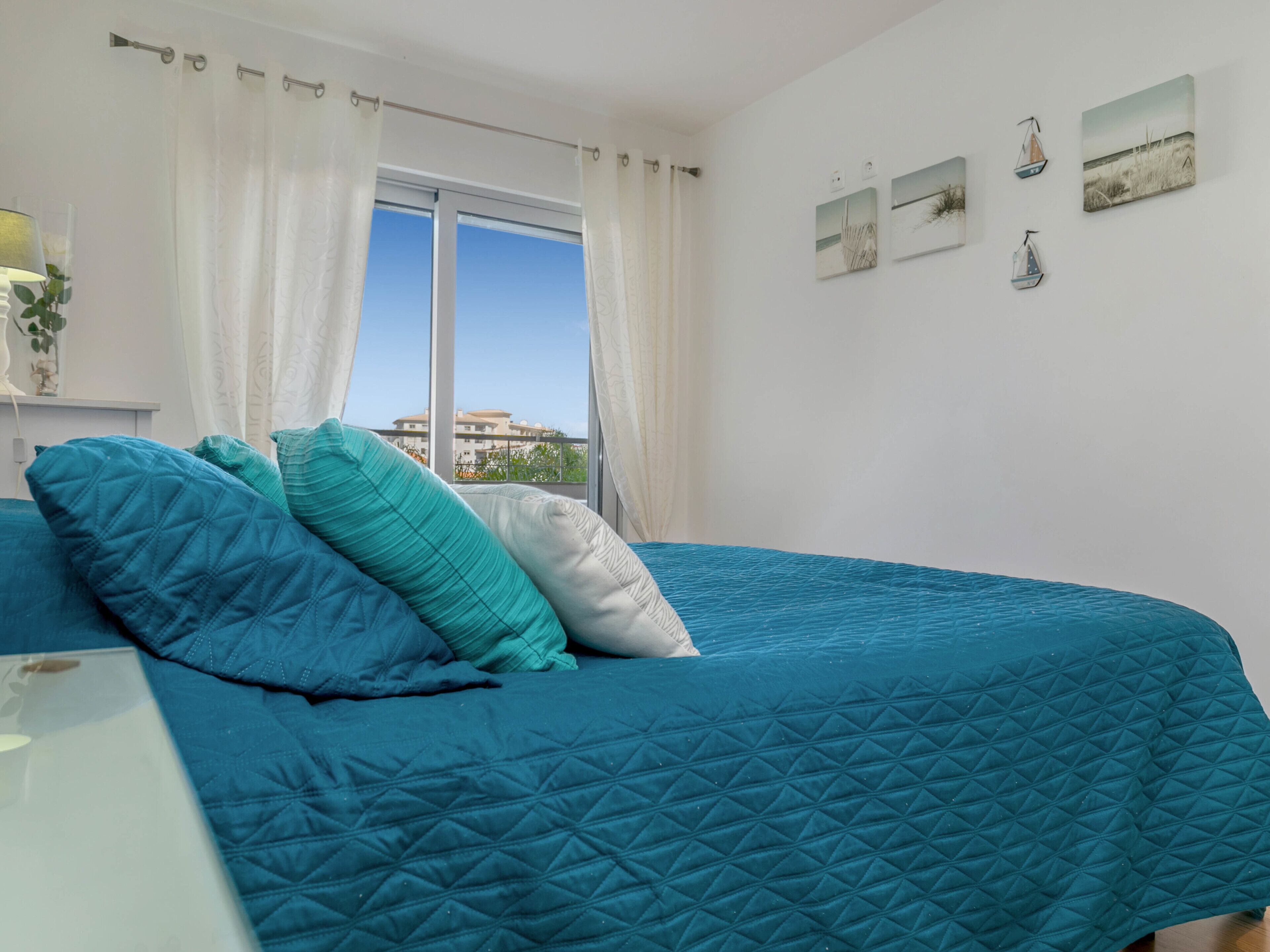 A bedroom with a large bed, white curtains, and a view of buildings through a window.