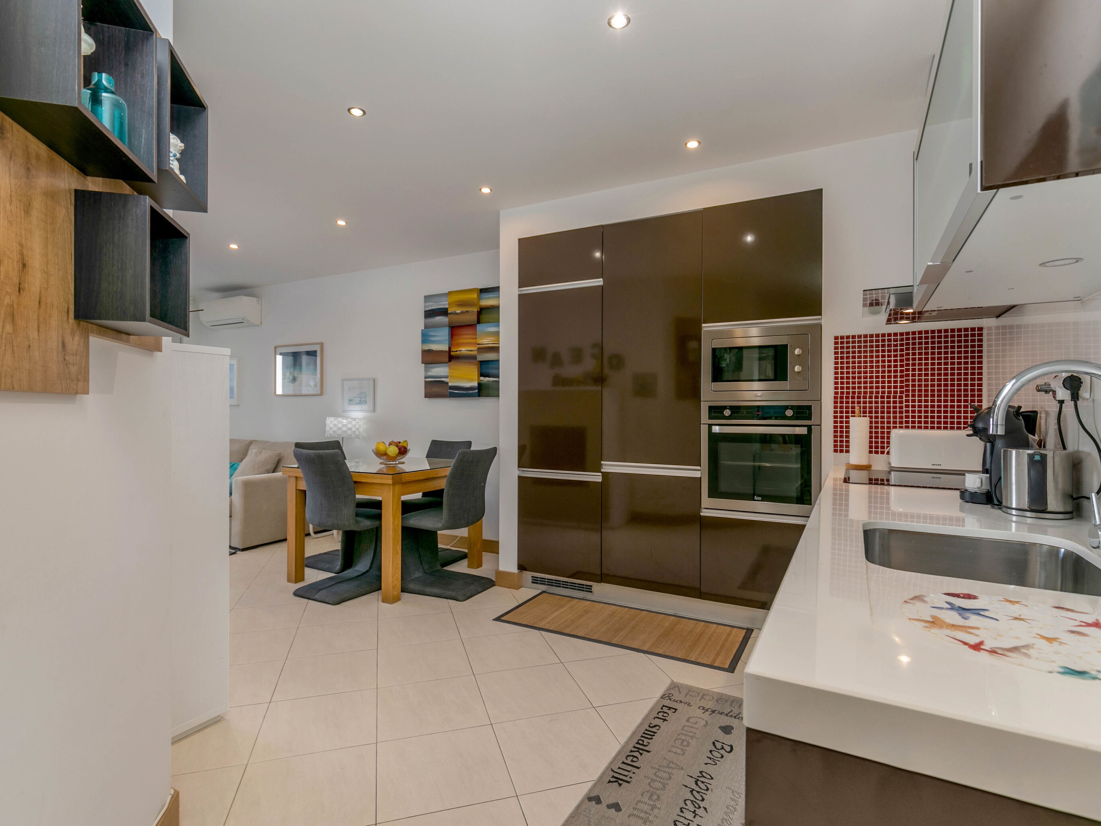 A modern kitchen with a dining area, featuring a built-in oven, a coffee machine, and a sink.
