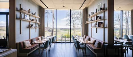 Un restaurante con mesas altas y bancos, ventanas amplias y estantes con diversos objetos.