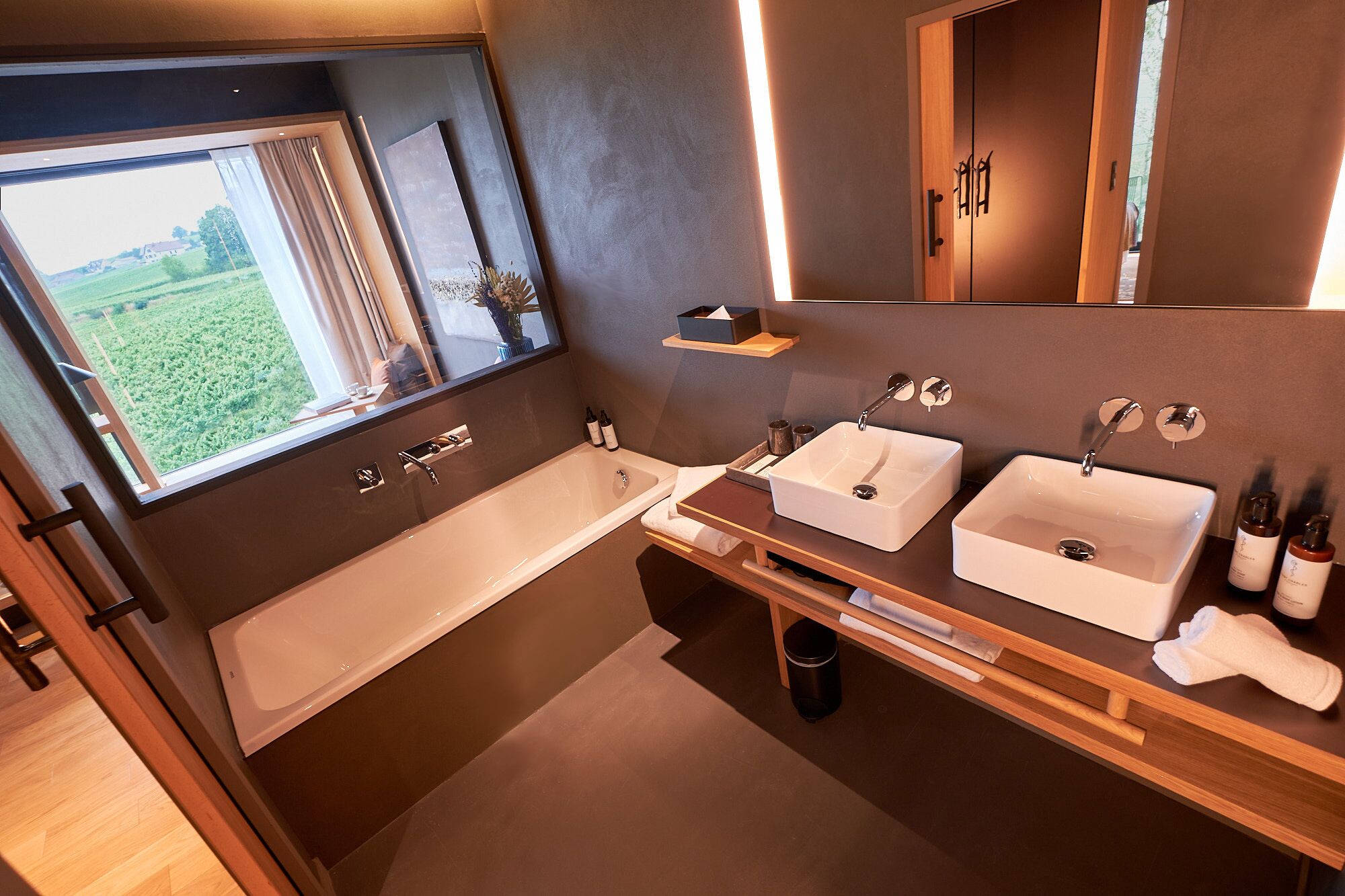 A modern bathroom with a large window, a bathtub, two sinks, and wooden shelves.