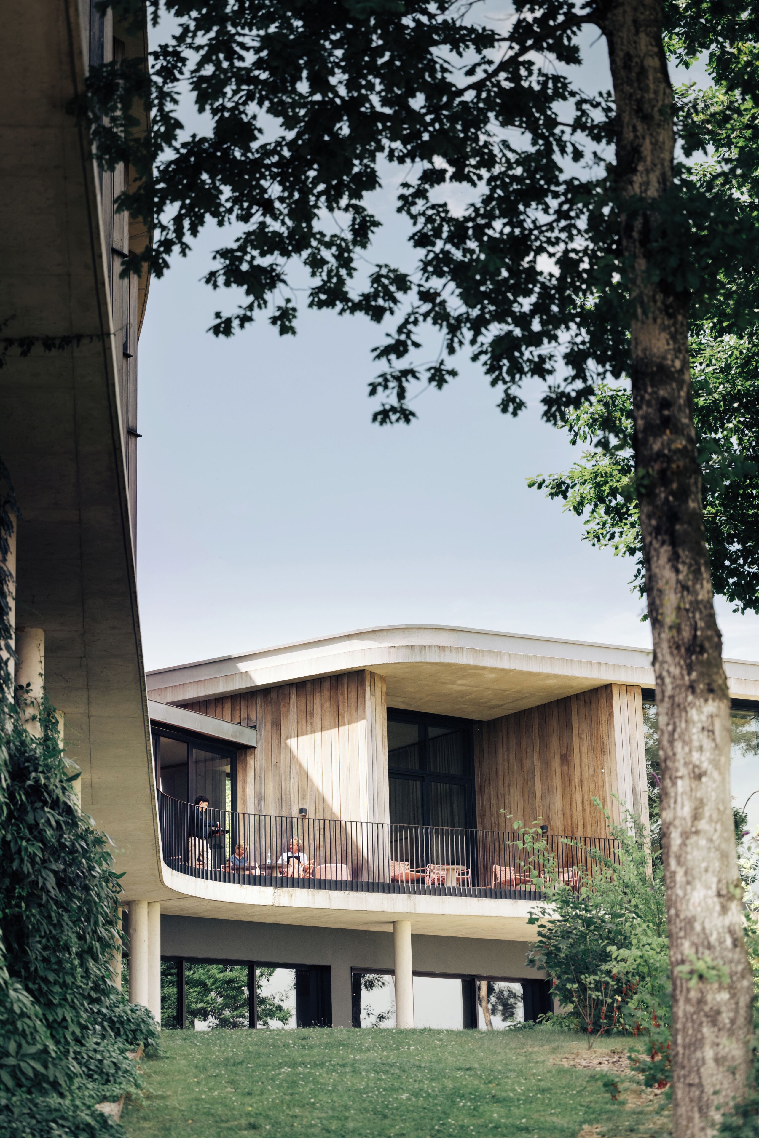 A modern building with a curved facade, a balcony with seating, and a grassy area in front.