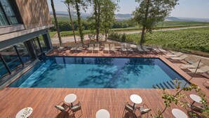 Ein moderner Poolbereich mit Liegestühlen und Tischen, umgeben von einer Holzterrasse mit Blick auf einen Weinberg.