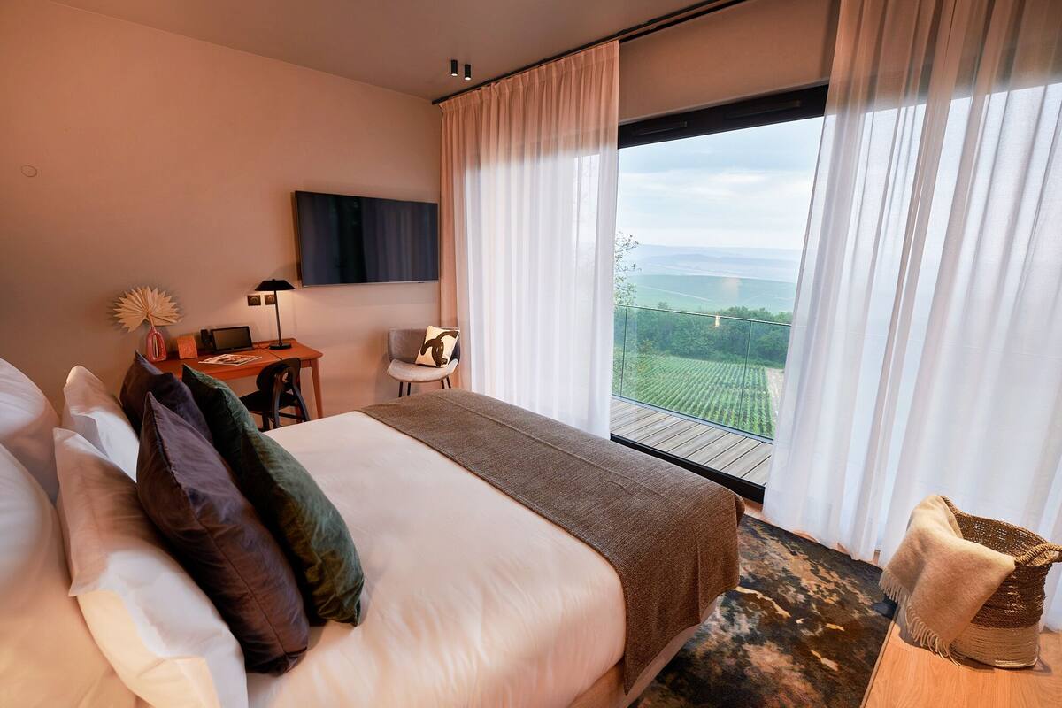 Hotelkamer met een groot bed, een bureau met een laptop, een stoel en uitzicht op een groen landschap door het raam.