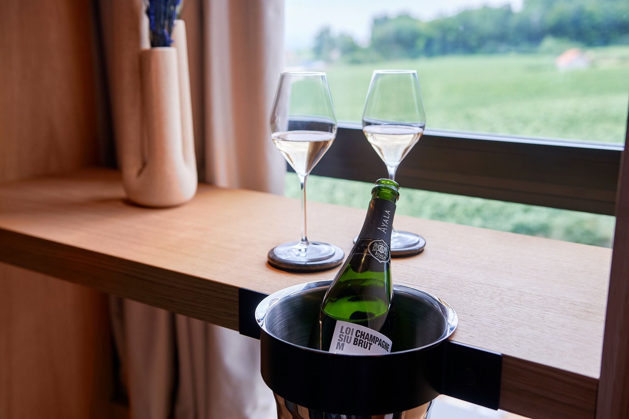 A bottle of champagne in a black bucket on a wooden ledge with two glasses of champagne.