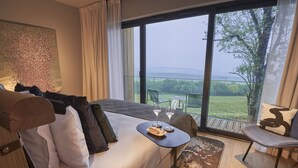 Ein Schlafzimmer mit einem großen Bett, einem kleinen Tisch mit Gläsern und Snacks, einem Sessel und Blick auf einen Garten und Berge durch große Fenster.