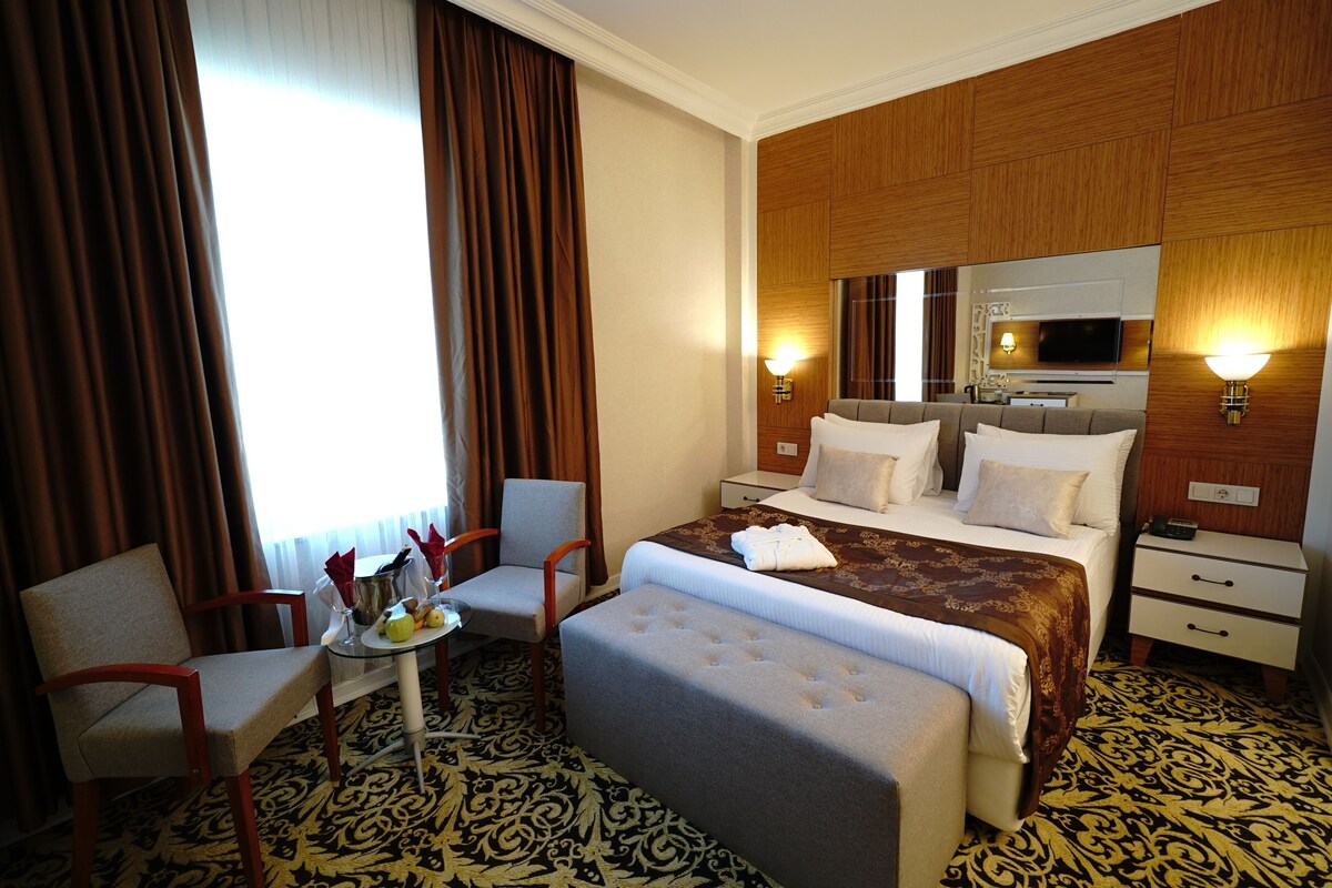 A hotel room with a bed, a chair, a small table with a lamp, a TV, and a large window with curtains.