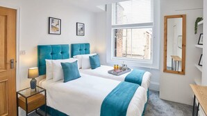 Ein Hotelzimmer mit einem großen Bett, einem Nachttisch mit Lampe, einem Fenster mit Jalousien, einem Spiegel und einer kleinen Pflanze auf einem Regal.