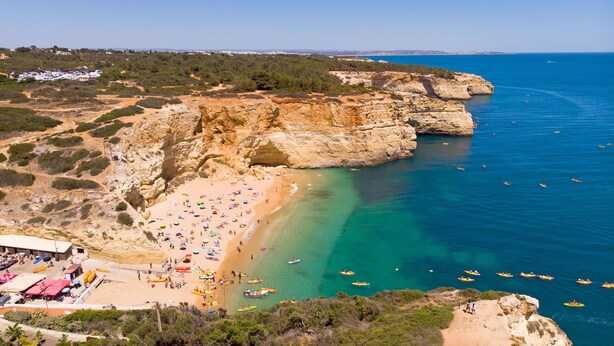 A coastal beach with clear waters, rocky cliffs, and a sandy shore.