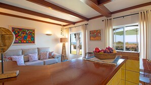 A spacious living area with a wooden ceiling, a large wooden table, a sofa with patterned cushions, and a view through a glass door to the outdoors.