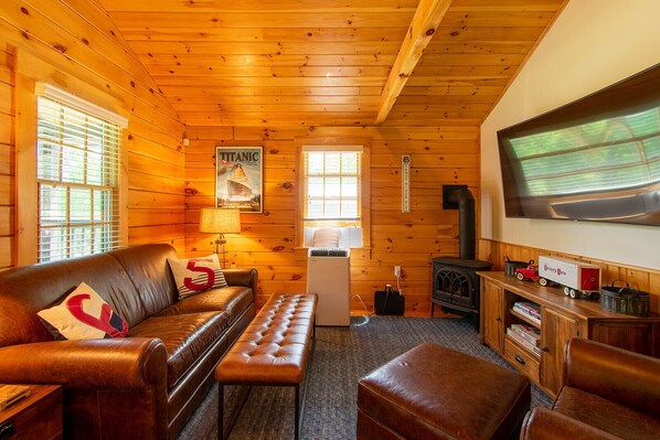 A cozy cabin interior with a leather sofa, a bench, a television, a wood-burning stove, and a sign indicating a scenic view of the Western River.