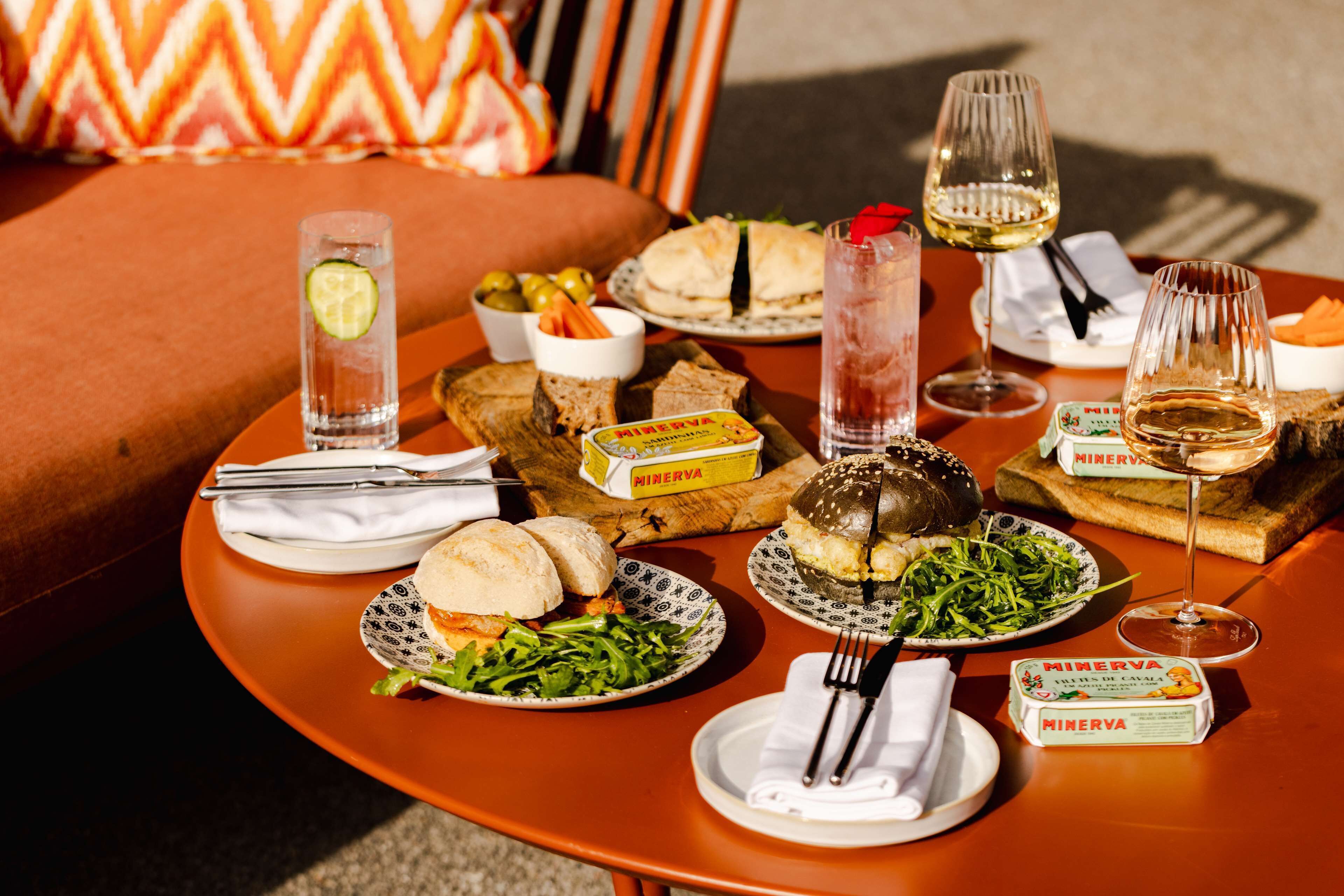 Une table dressée avec divers plats, notamment des sandwichs, des salades et des boissons, sur une table d’extérieur, avec un coussin en arrière-plan.