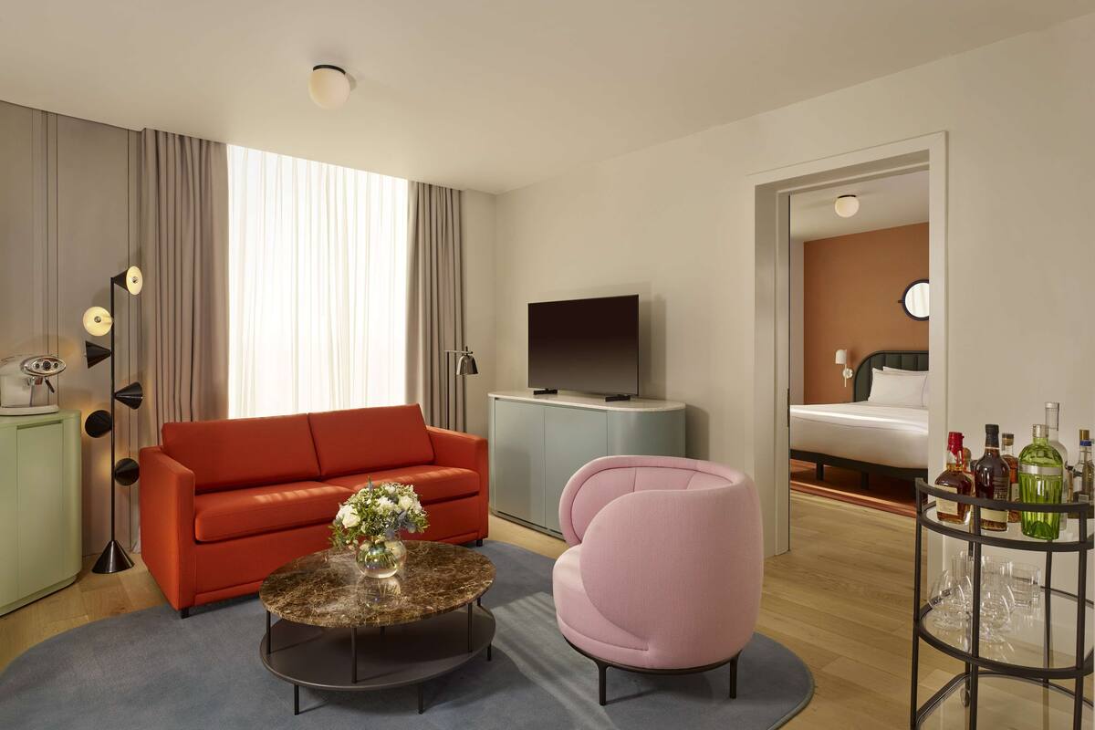 Une chambre d’hôtel moderne avec un canapé rouge, un fauteuil rose, une table basse ronde ornée d’un vase, un téléviseur à écran plat, un minibar et un meuble vitré contenant des bouteilles.