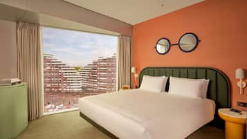 Habitación de hotel con una cama grande, vistas a edificios altos, un espejo redondo en la pared y una mesita de noche con una lámpara.