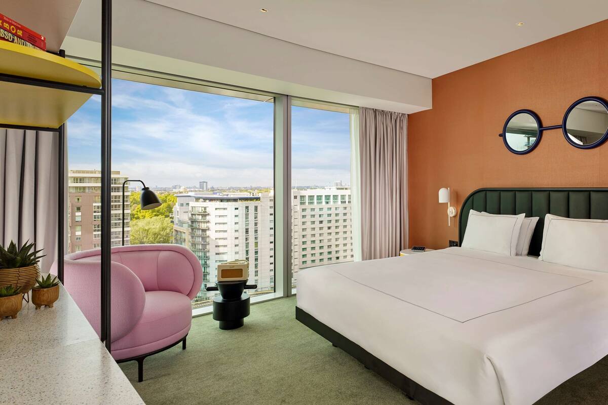 Une chambre d’hôtel moderne avec un grand lit, un fauteuil rose, une petite table et une vue sur le paysage urbain grâce à de grandes fenêtres.