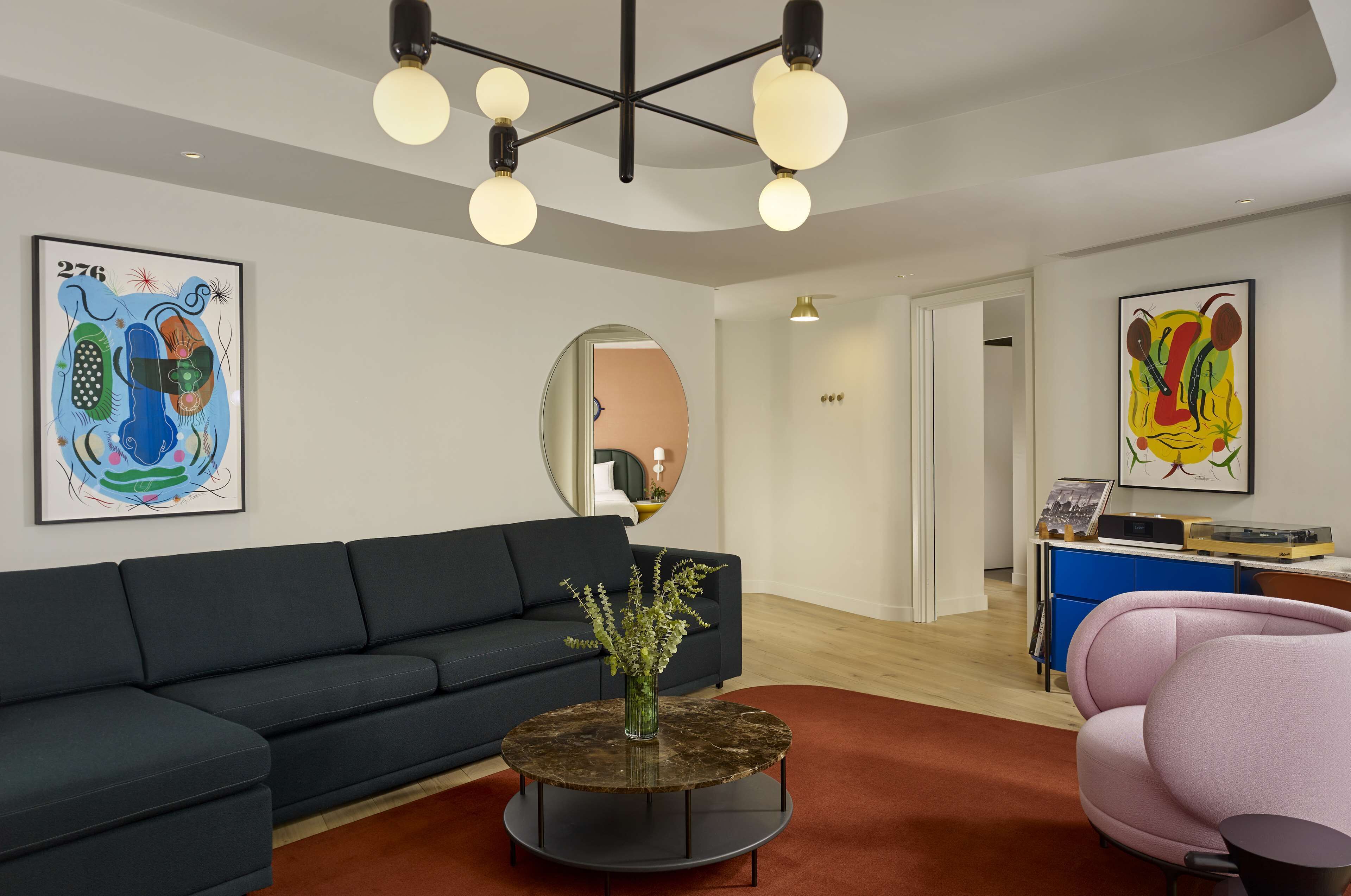 A modern living room with a dark sofa, a round coffee table with a vase, and a pink chair. There are abstract paintings on the walls and a ceiling light fixture.
