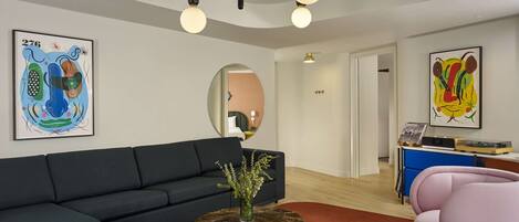 A modern living room with a dark sofa, a round coffee table with a vase, and a pink chair. There are abstract paintings on the walls and a ceiling light fixture.