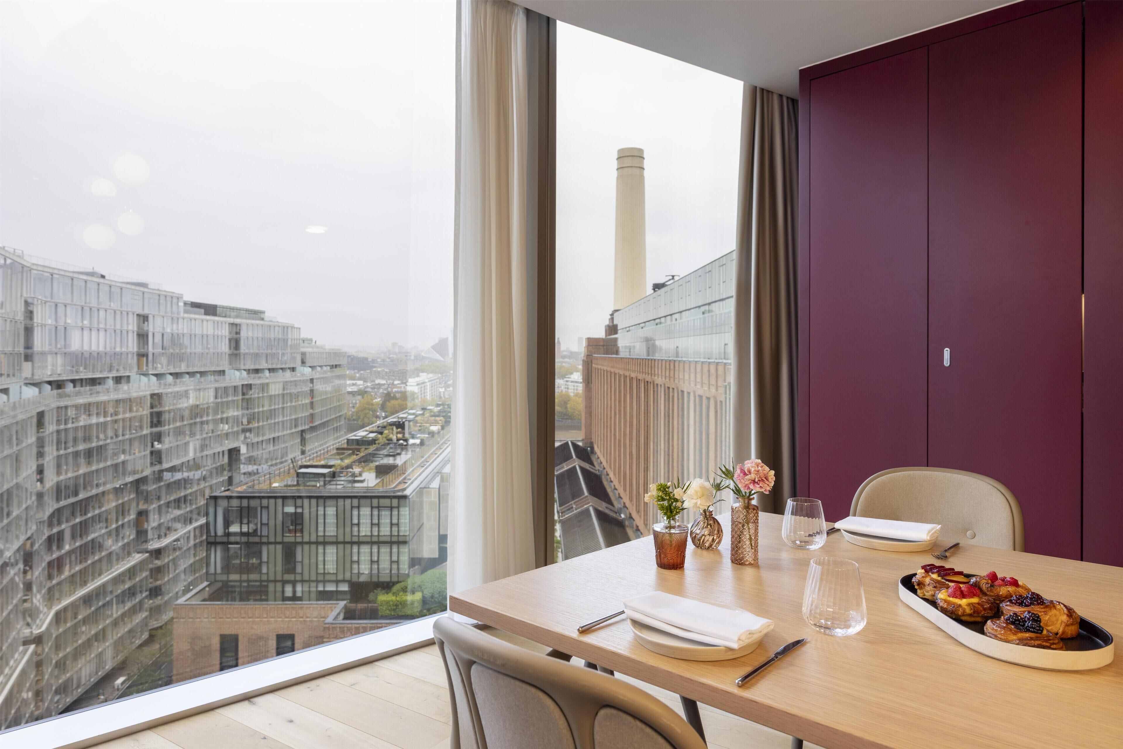 Een moderne hotelkamer met een groot raam met uitzicht op de stad, een eettafel met ontbijt en een paarse garderobe.