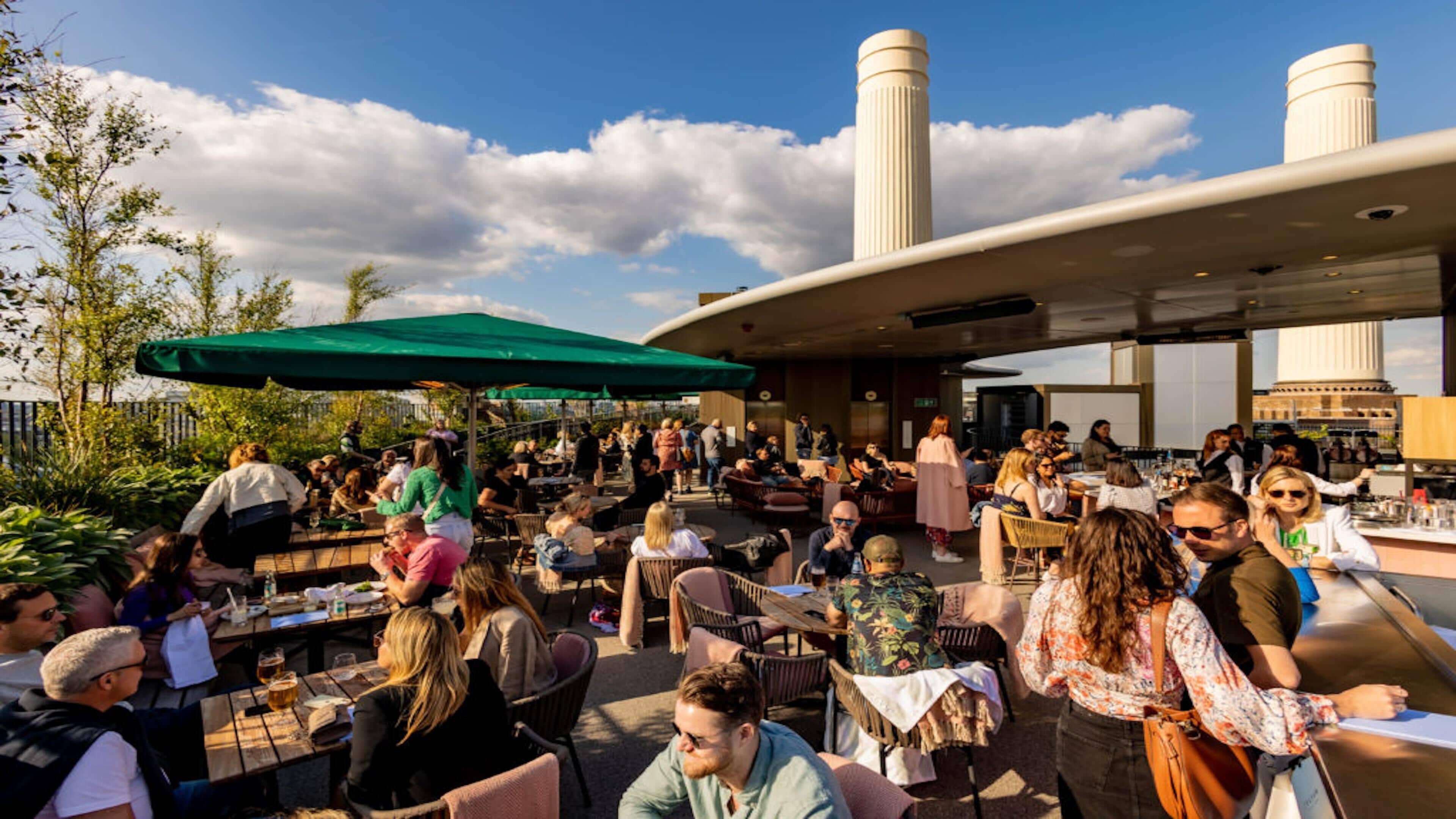 An outdoor dining area with tables and chairs, a green umbrella, and a modern building with cylindrical structures in the background.