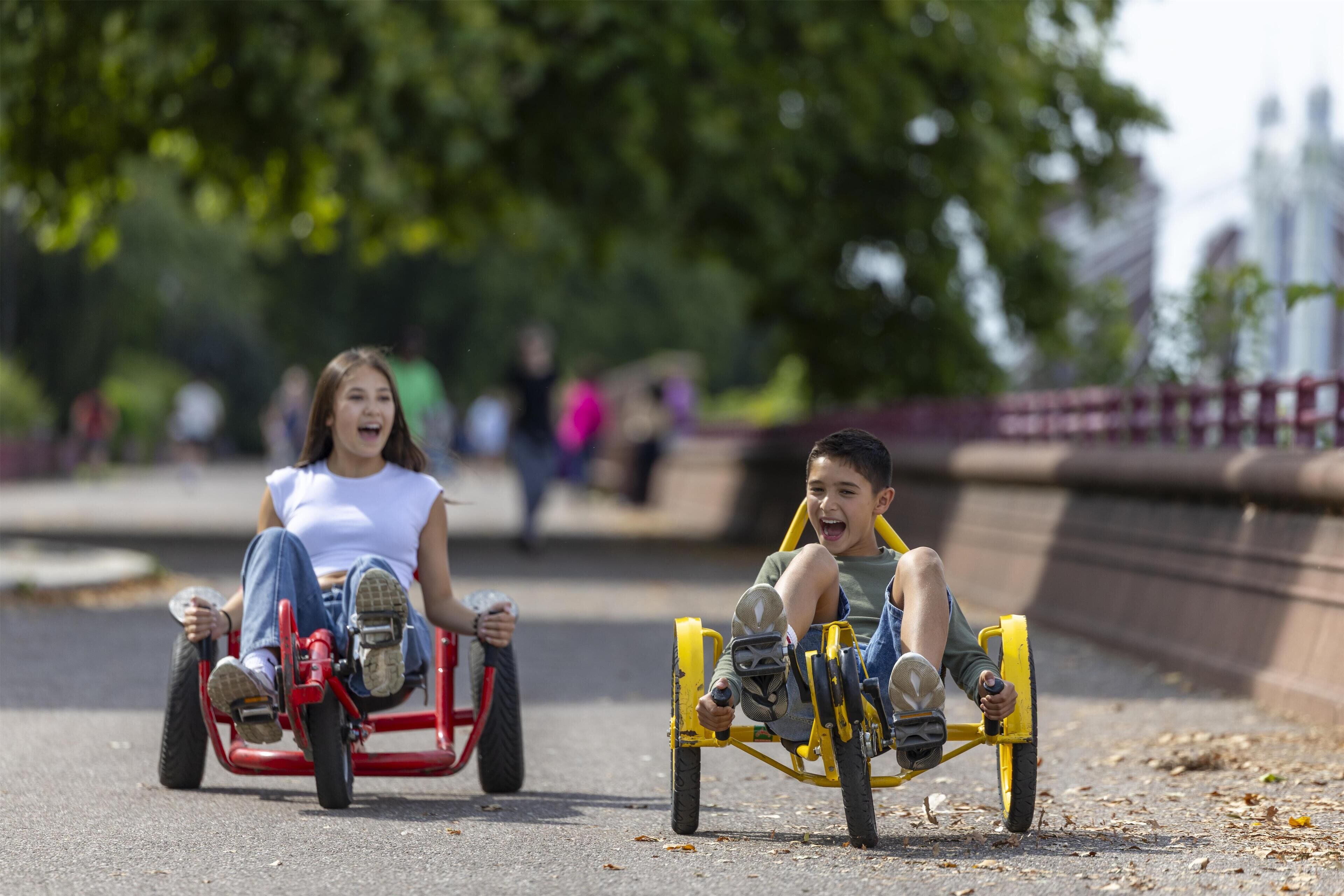 Two children on a red and yellow tricycle on a paved path.