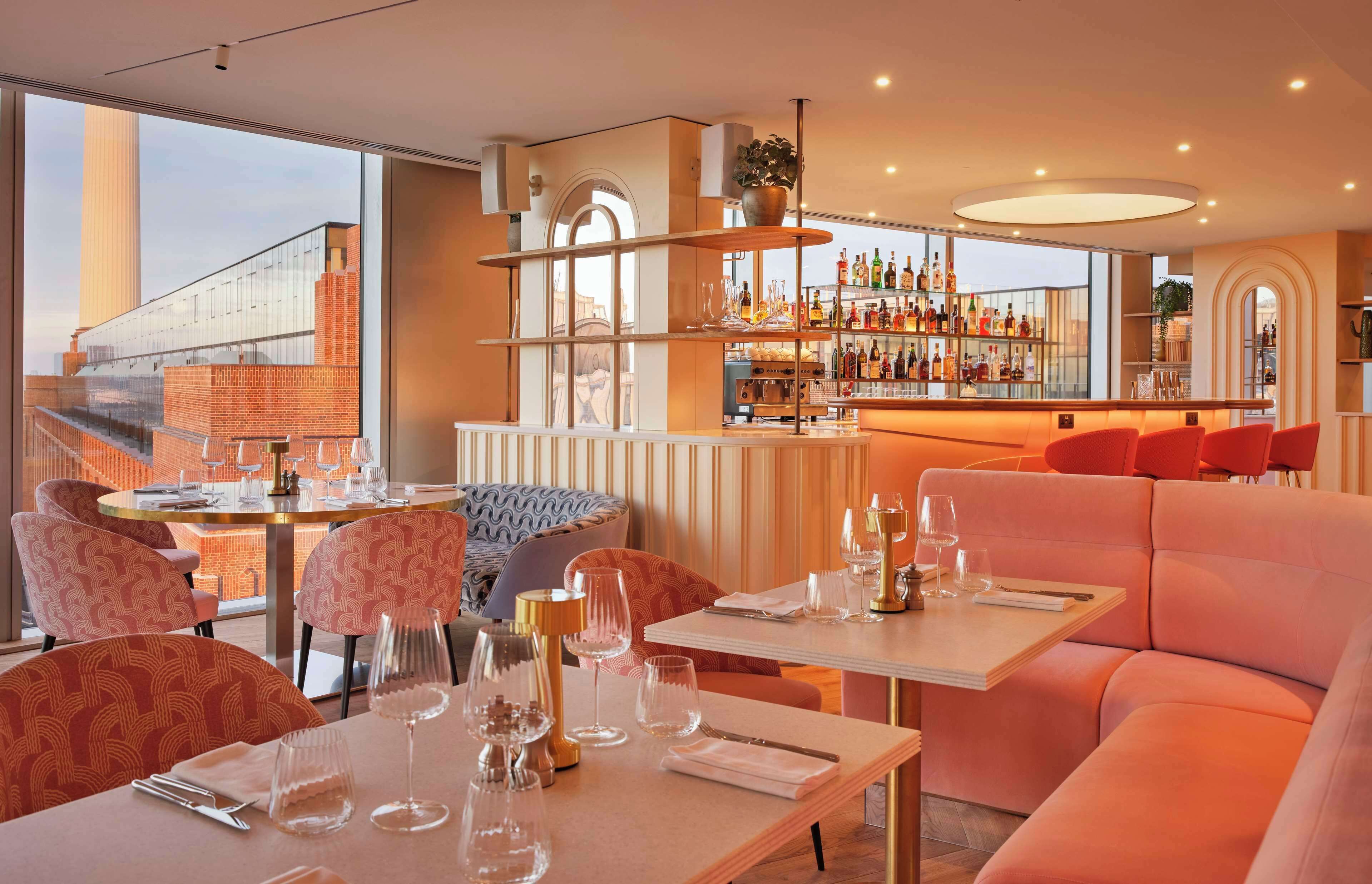 A modern bar area with a glass wall, a variety of bottles, and a seating area with tables and chairs.