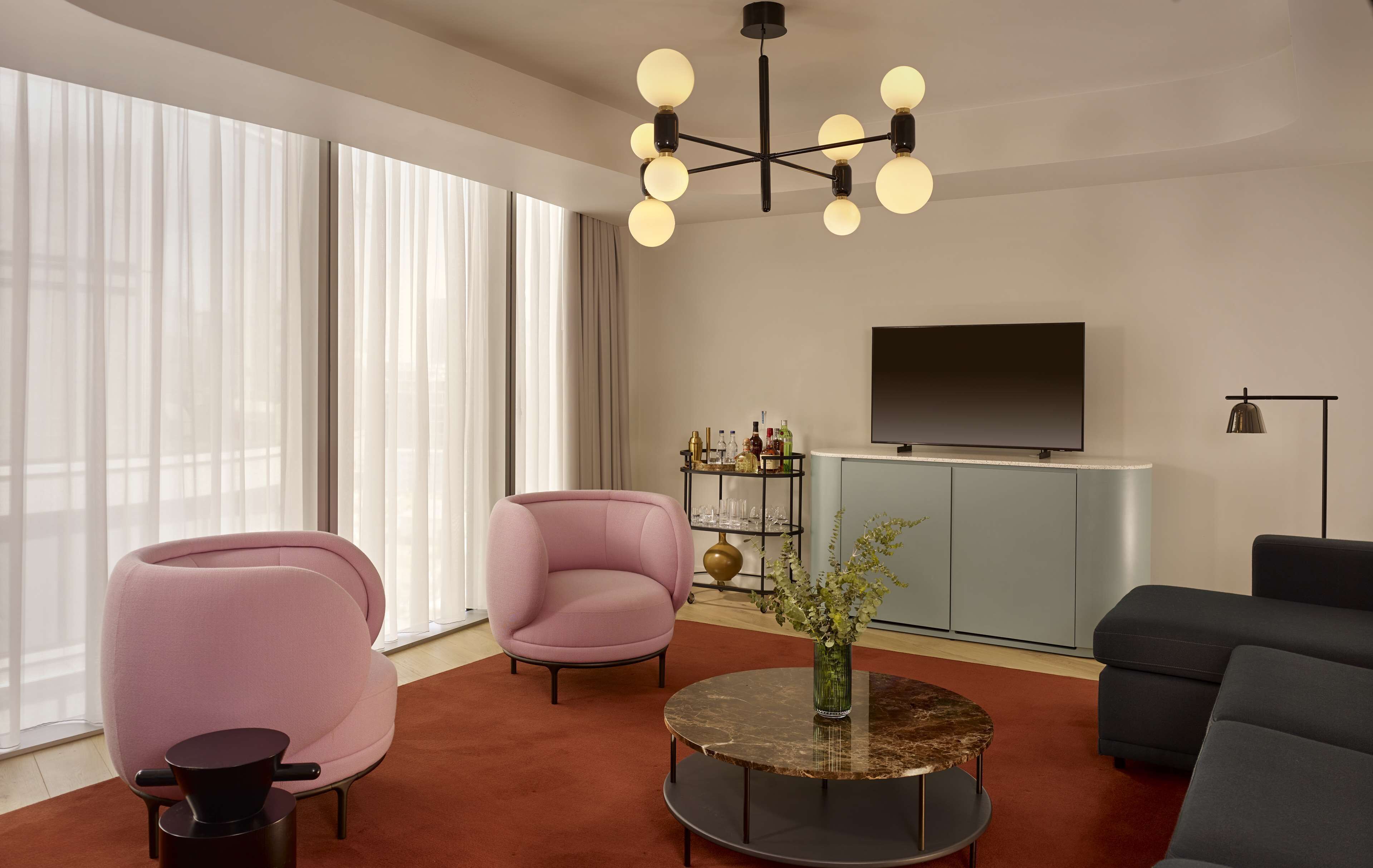 A modern living room with a pink armchair, a round coffee table with a vase, and a grey sofa. A flat-screen TV is mounted on the wall, and there is a bookshelf with various items.