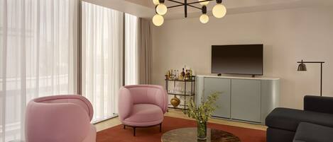 A modern living room with a pink armchair, a round coffee table with a vase, and a grey sofa. A flat-screen TV is mounted on the wall, and there is a bookshelf with various items.