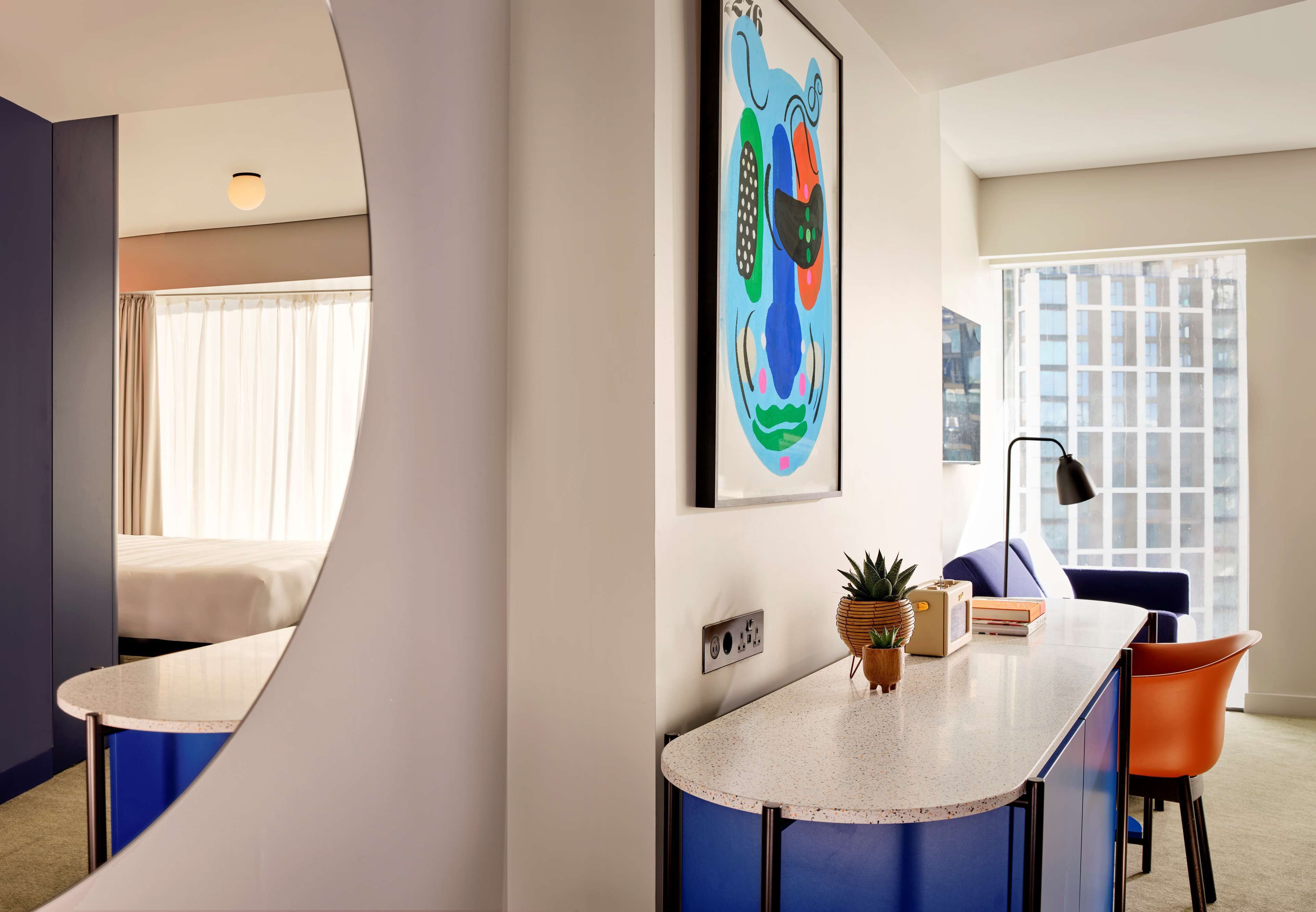 A modern hotel room with a curved counter, a bed, a large window, and a colorful abstract painting on the wall.