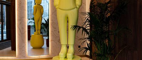 En gul statue af en person, der står på en piedestal i en lobby med marmorvægge og et stort spejl, der reflekterer statuen.