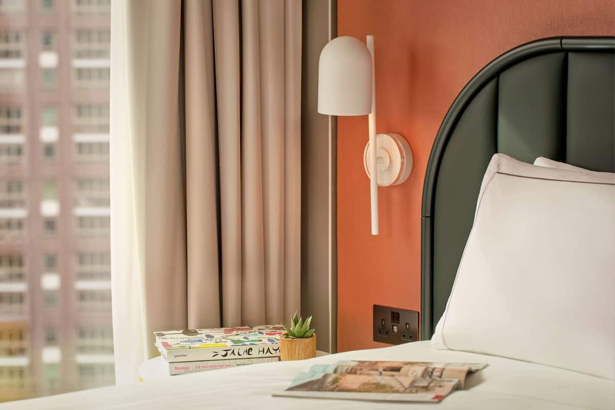 Une chambre d’hôtel avec un lit, une lampe de chevet, une petite plante et des livres sur la table de chevet.