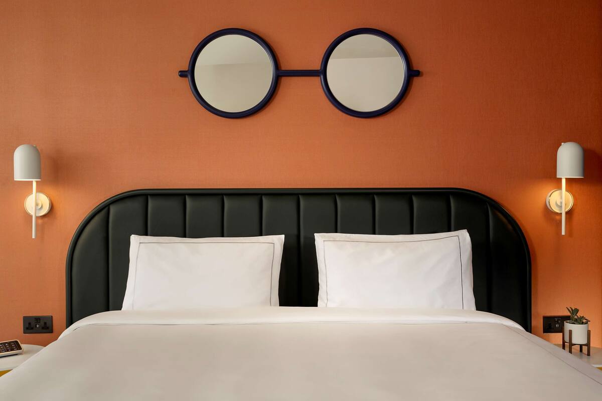Une chambre d’hôtel avec une tête de lit noire, deux oreillers blancs, un miroir rond et des lampes fixées au mur.