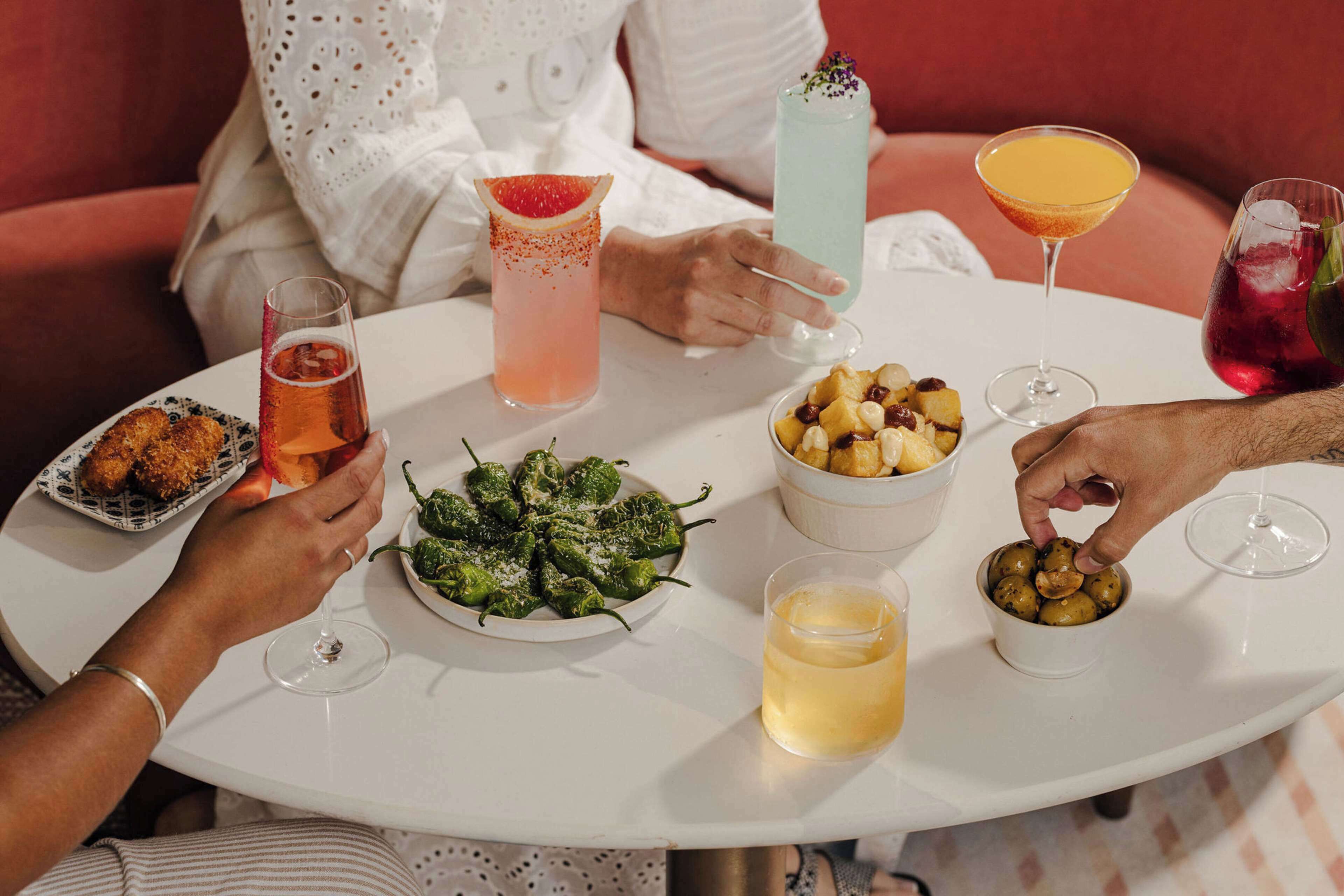 A round table with various drinks and food items, including a plate of green peppers, a bowl of fruit, and a glass of beer.
