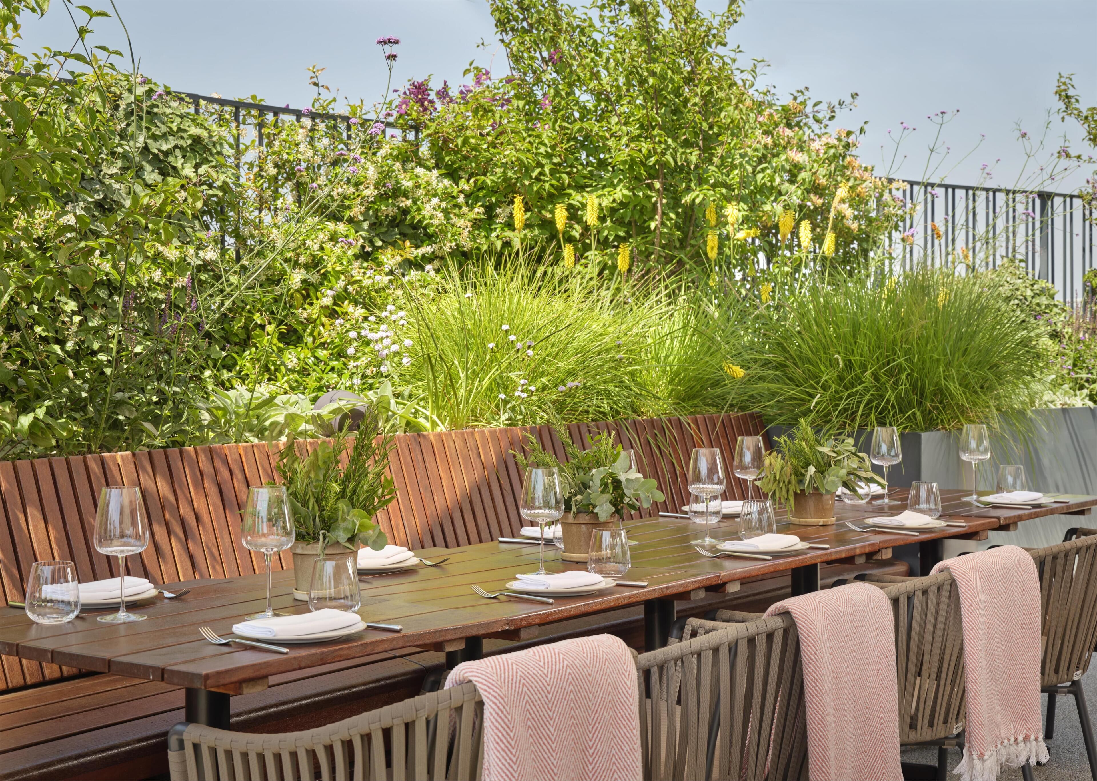 Un espace de restauration en plein air avec une longue table en bois dressée pour six personnes, entourée de chaises recouvertes de plaids roses.