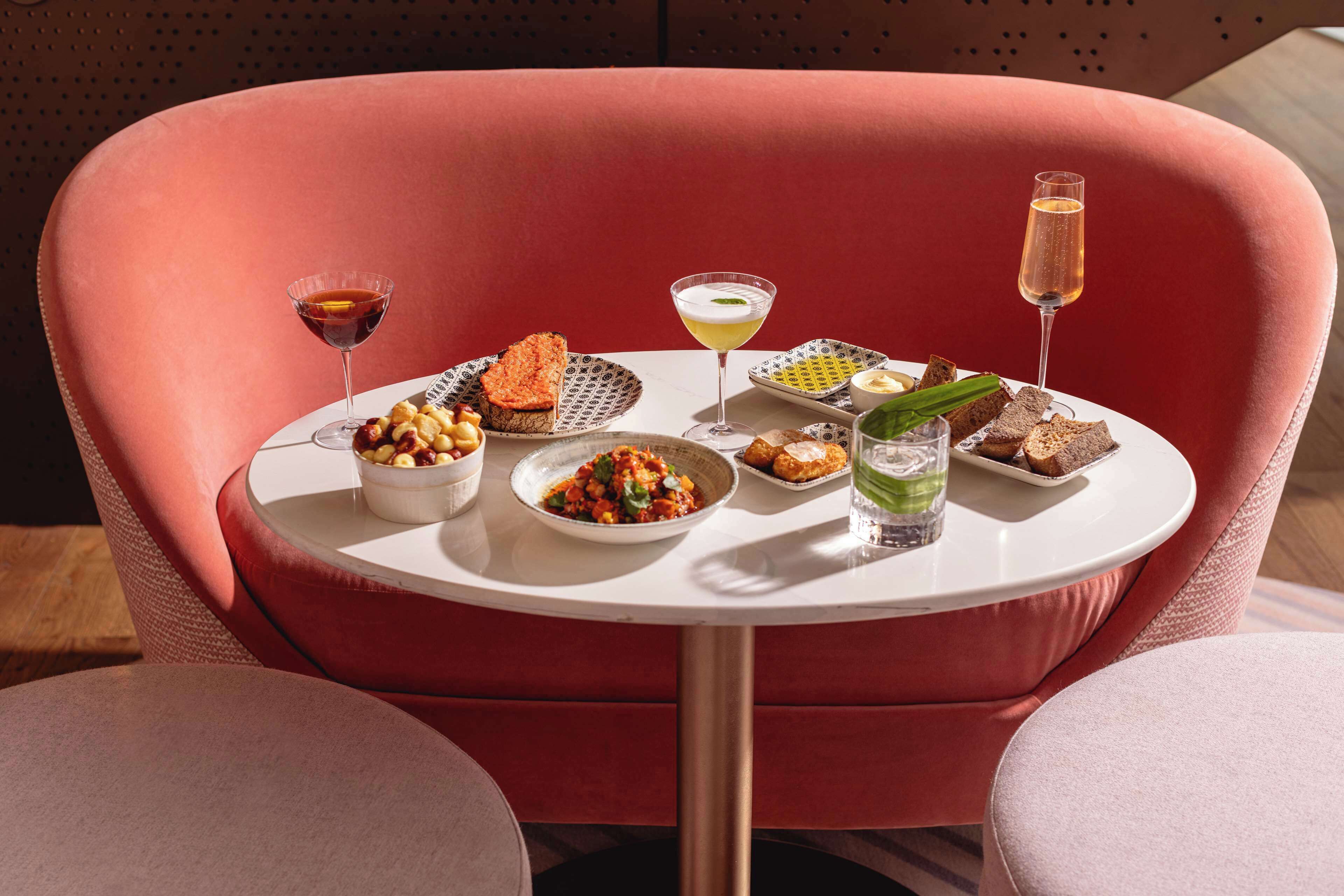 A table with a variety of dishes and drinks, set on a round table in a room with a red upholstered chair.