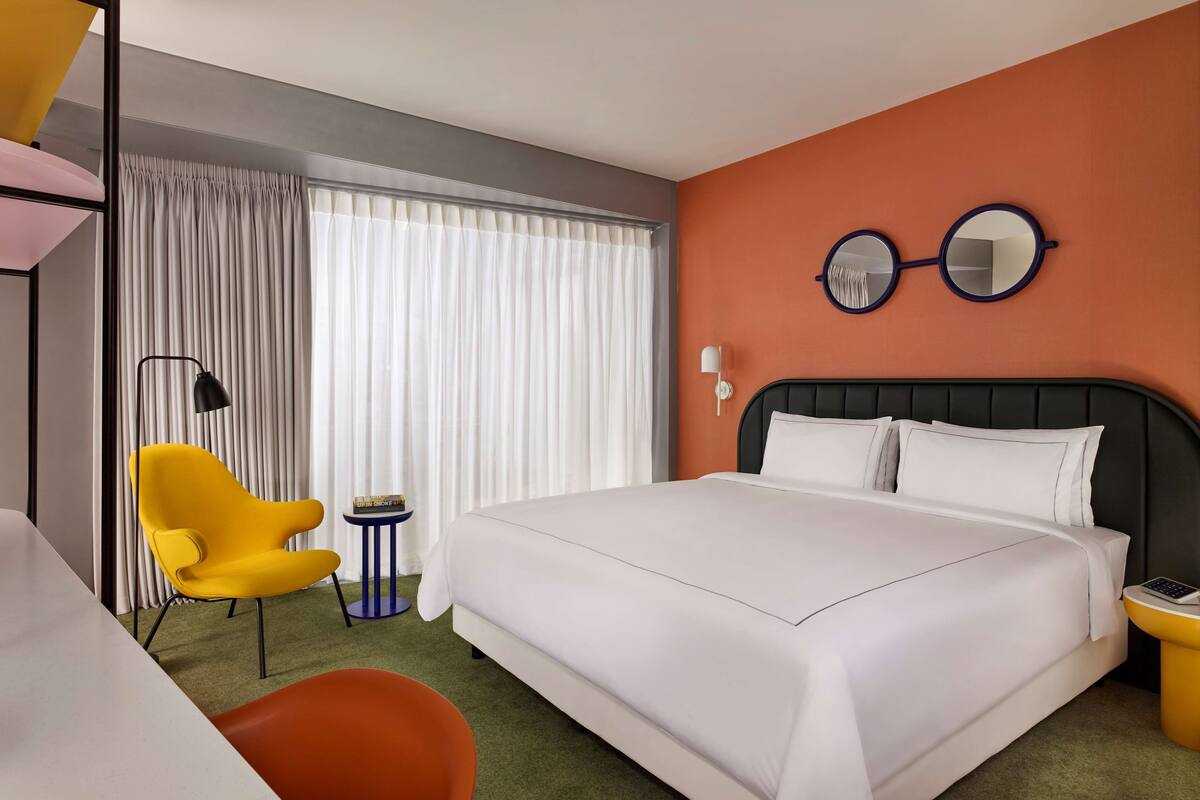 Une chambre d’hôtel avec un grand lit, une chaise jaune, une petite table ronde et un miroir au mur.