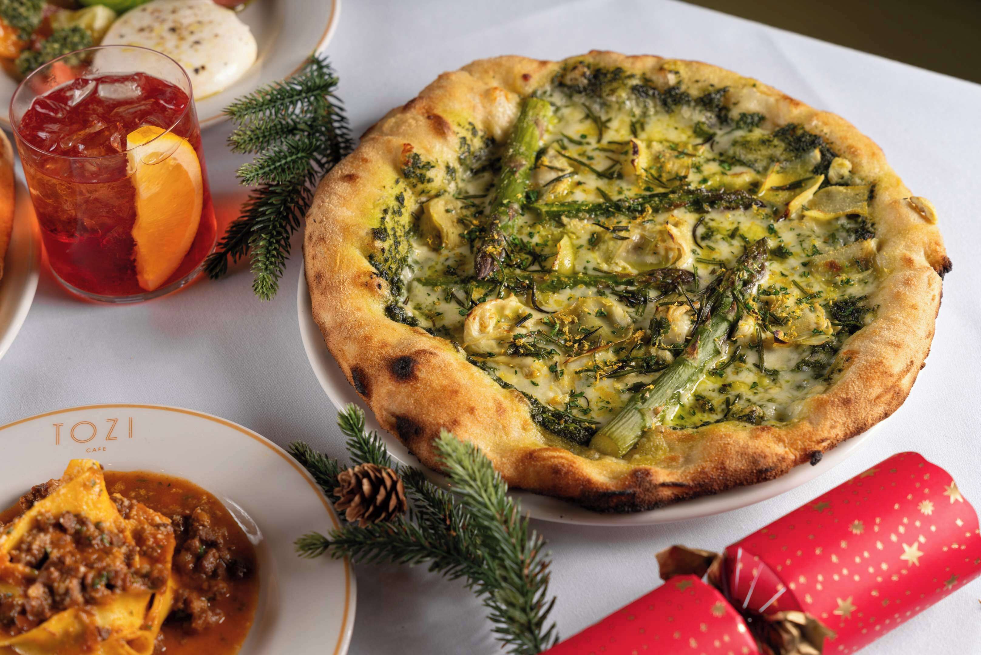 Una pizza con broccoli e formaggio su un piatto, un bicchiere di bevanda rossa e un piatto di ravioli con salsa.