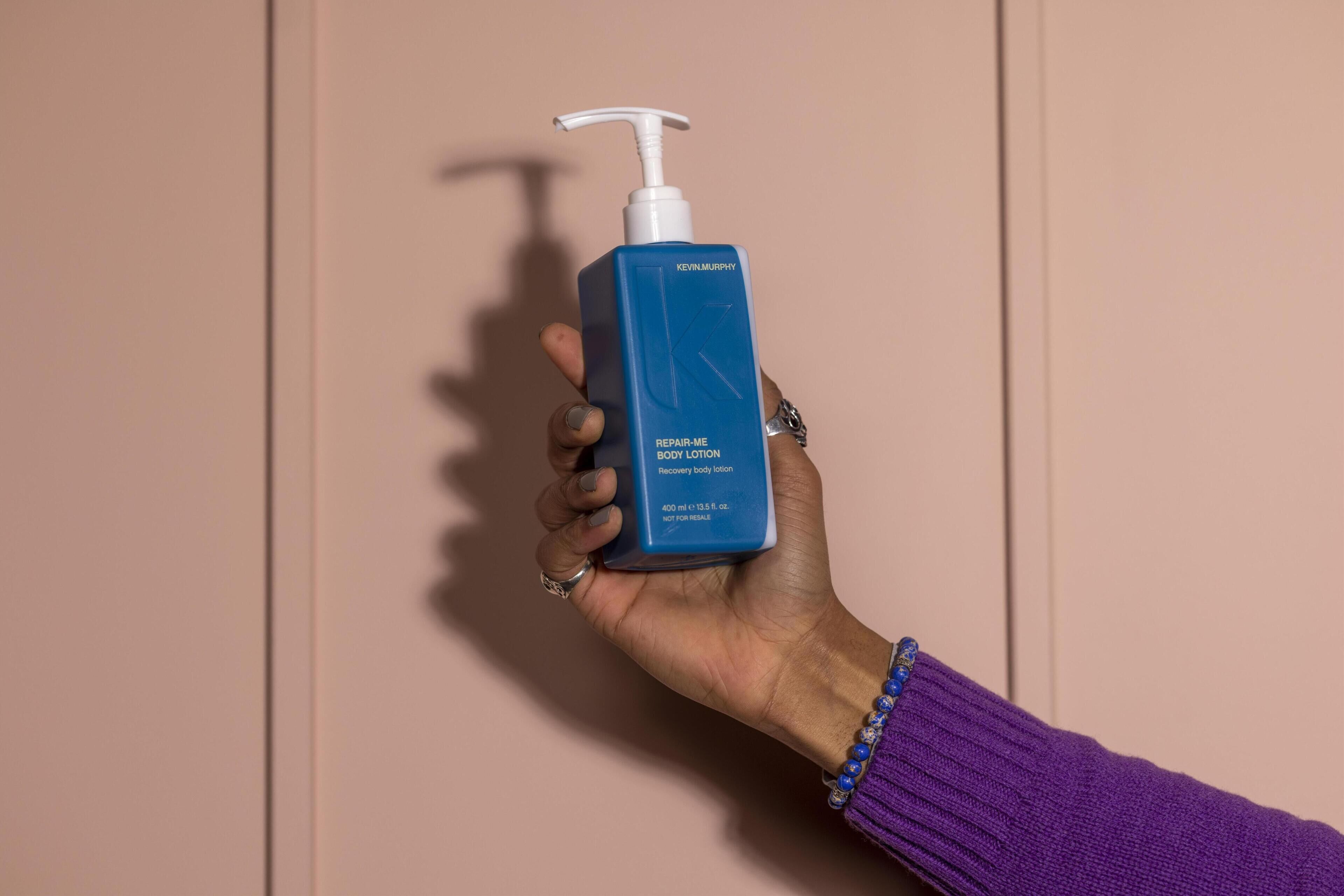 A hand holding a blue bottle of Kevin Murphy Repair-Me Body Lotion.
