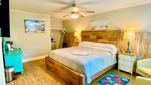 A bedroom with a wooden bed, a ceiling fan, a nightstand with a lamp, a chair, and a small table with a lamp.