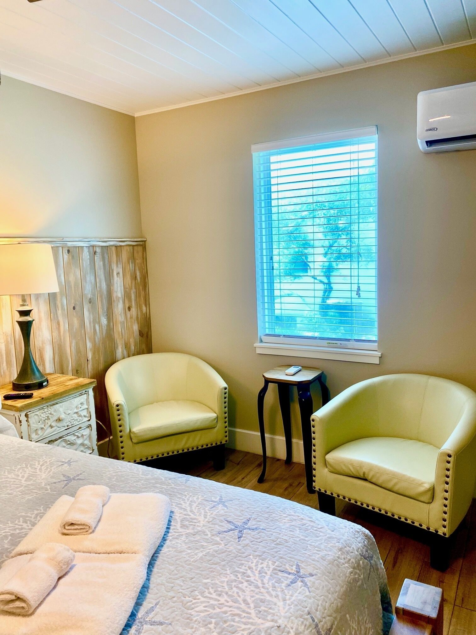A room with a bed, two chairs, a small table, a lamp, and a window with blinds.