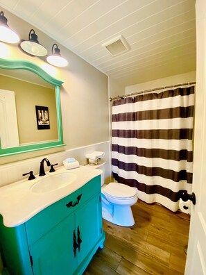 A bathroom with a turquoise vanity, a white sink, a black faucet, a white toilet, and a striped shower curtain. - Coast Oaks Cottage 6 1 Bedroom Cottage by RedAwning (Rockport)