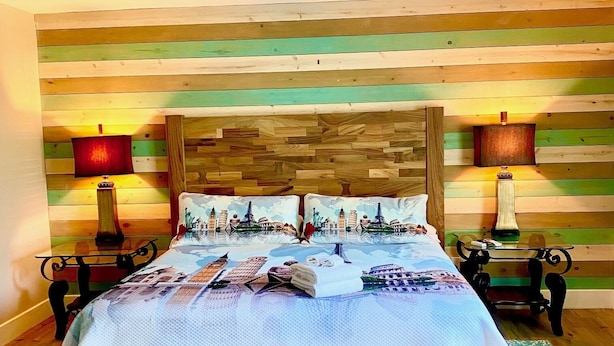 En säng med träram och ett sängöverkast med motiv av en stadsbild, två sänglampor och en takfläkt i ett rum med väggar klädda med träpaneler.