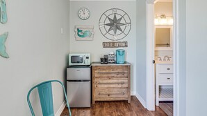 Eine kleine Küche mit Kühlschrank, Mikrowelle, Kaffeemaschine und einem Holzkästen. Eine Uhr und Wanddekoration sind zu sehen.