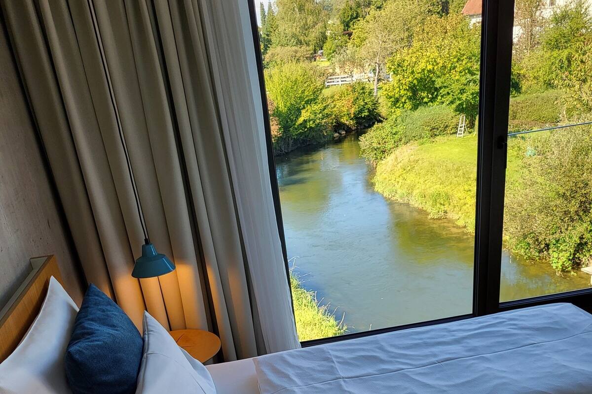 Ett hotellrum med en säng, en lampa och utsikt över en flod och grönområden.