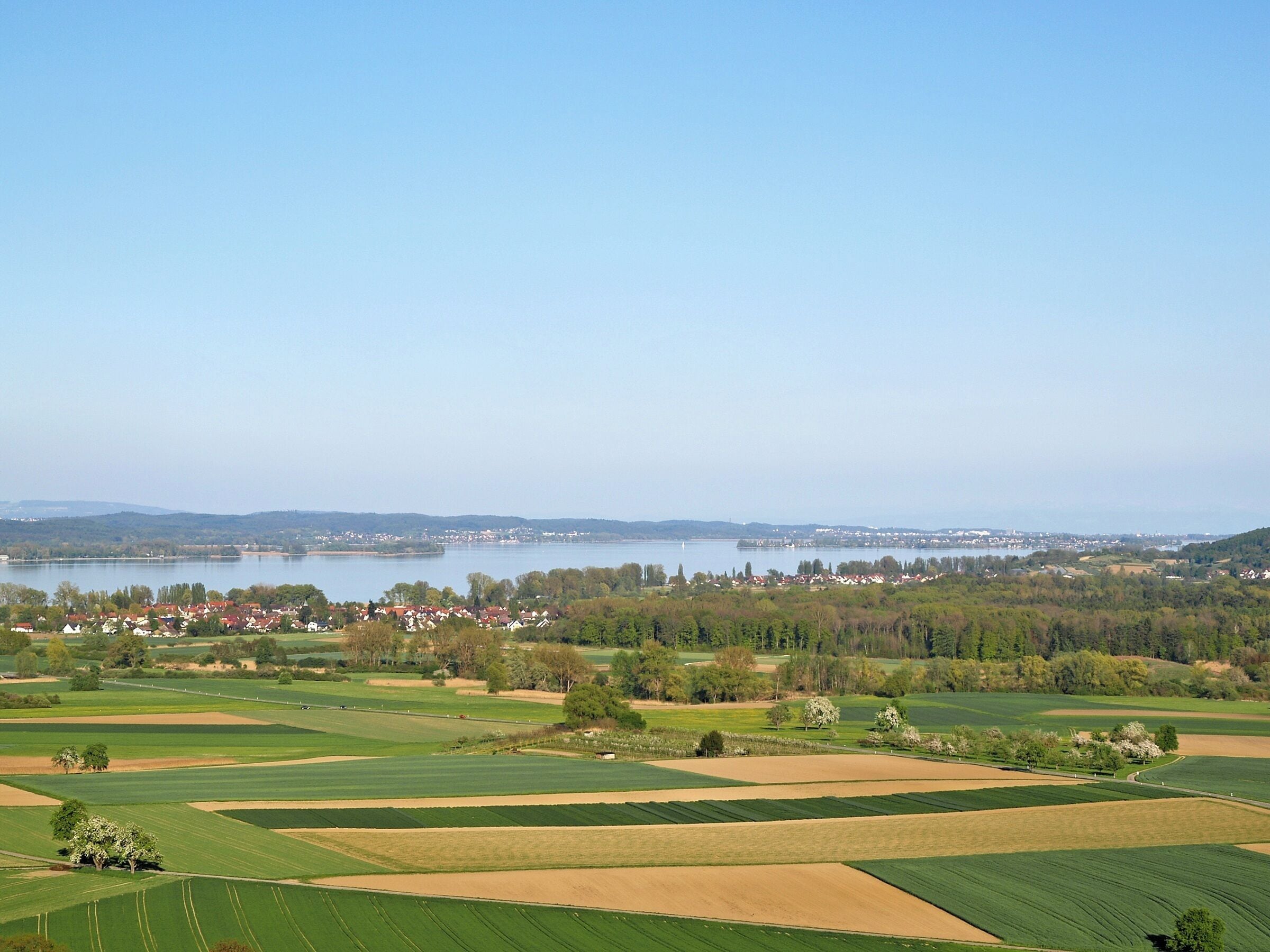 Een panoramisch uitzicht over een landelijk landschap met velden, een meer en een klein stadje in de verte.