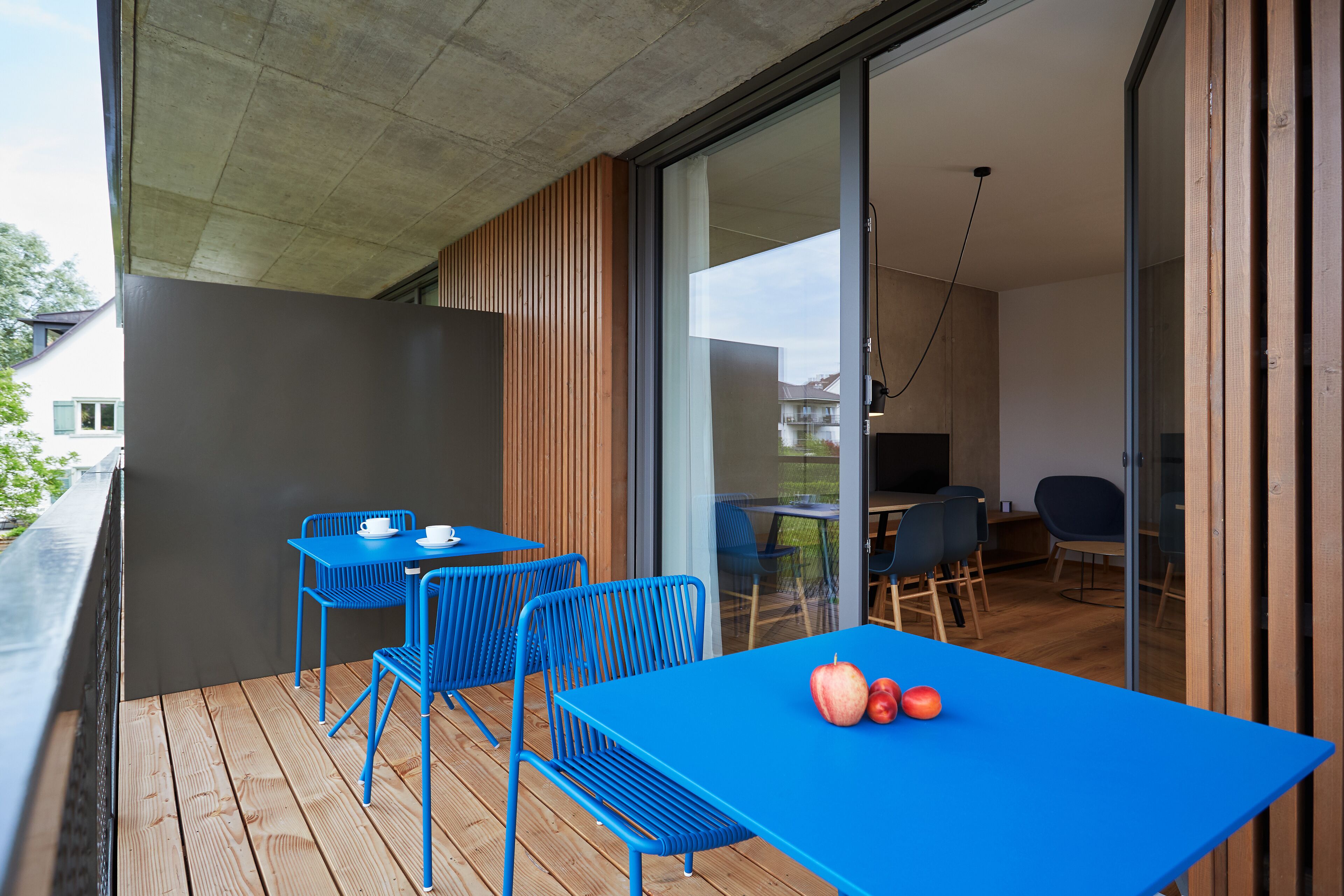 A balcony with blue metal chairs and a table, a wooden deck, and a sliding glass door leading to an interior space with a dining area.