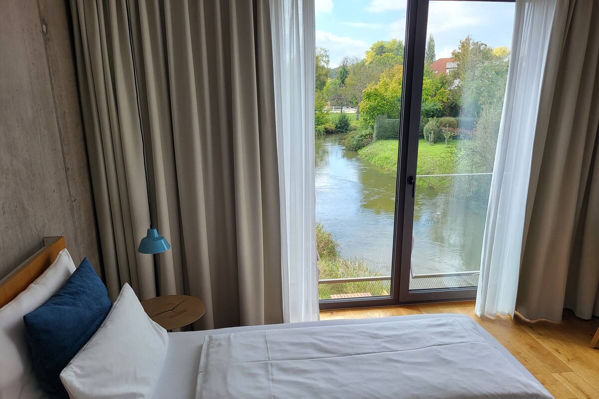 Ett sovrum med en säng, gardiner, ett nattduksbord, en lampa och utsikt över en flod och grönområden utanför.