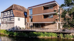 Un edificio moderno junto a una estructura tradicional de entramado de madera, con un paseo fluvial y una zona de estar al aire libre.